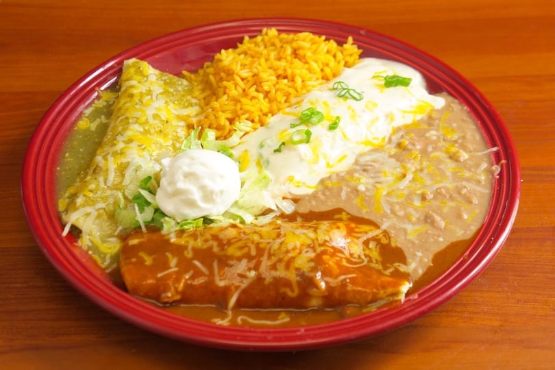 A red plate holds two burritos, rice, and refried beans, garnished with sour cream and lettuce.