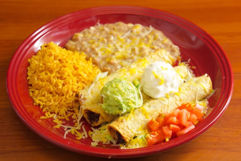 A red plate holds three enchiladas topped with guacamole and sour cream, alongside rice and refried beans.
