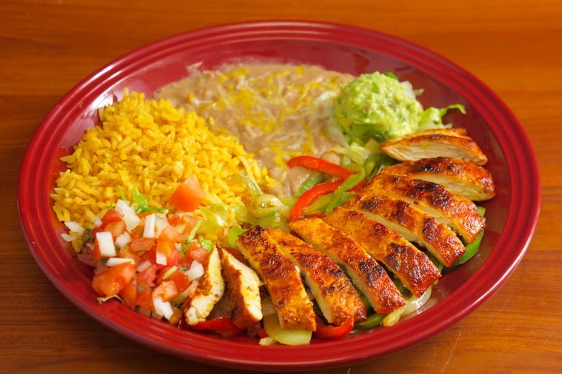 Grilled chicken fajitas with rice, refried beans, guacamole, and pico de gallo on a red plate.