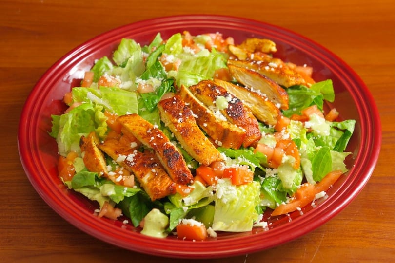 Grilled chicken salad with lettuce, diced tomatoes, crumbled cheese, and green dressing in a red bowl.